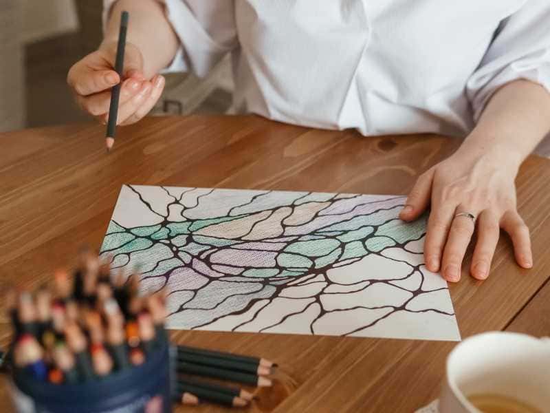 Eine Person hält einen Buntstift und zeichnet auf einem Blatt Papier mit einem neurographischen Muster. Auf dem Tisch sind Farbstifte und eine Tasse sichtbar.