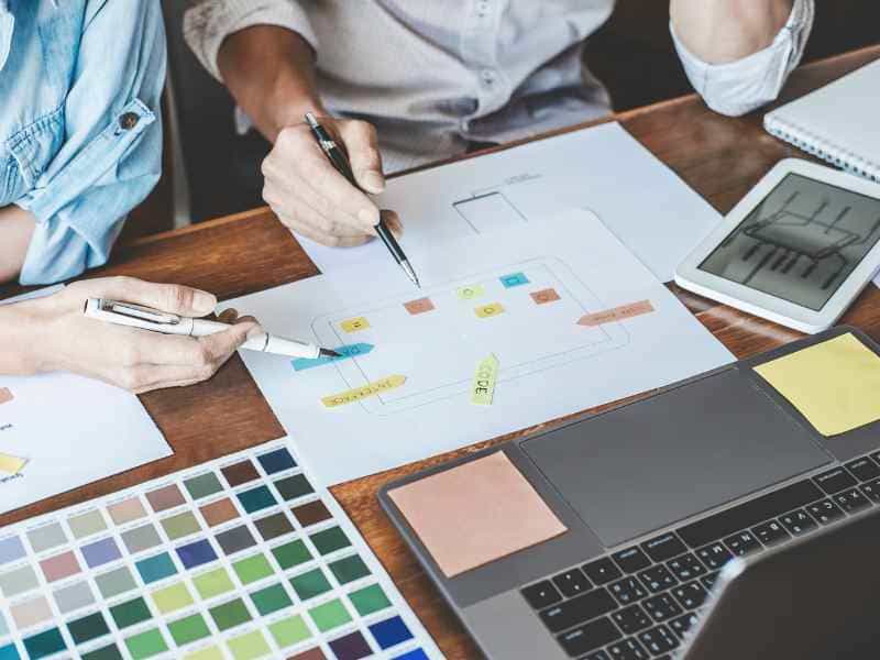 Zwei Personen planen eine Website: Skizzen mit Post-its, Farbpalette, Laptop und Tablet – Symbol für Website-Konzept, Design und WordPress-Umsetzung.