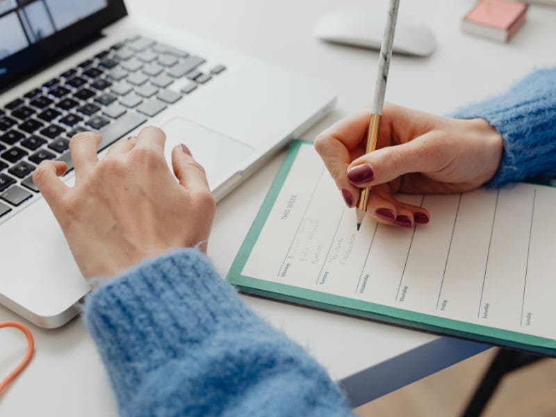Start 3 Frau schreibt am Laptop in einen Planer und sortiert Gedanken für den Start in die Selbstständigkeit